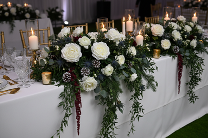 Artificial white silk open roses styled in an elegant wedding top table centrepiece with white roses, green foliage, pinecones and tall glass candle holders with lit candles in a gold and white wedding venue
