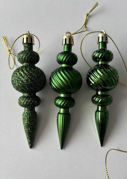 Close up of three green Christmas spindle baubles side by side showing glittered, metallic and matte finishes with ribbed spherical tops, beaded middles and elongated pointed drop bases with gold hanging strings on a plain white background