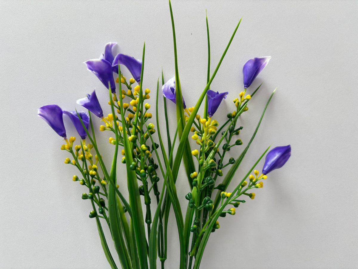 Full spread shot of an artificial purple small calla lily foliage bunch showing five individual wired stems fanning out with small purple calla lily flower heads, tall thin green grass stems and small bright yellow berry pick stems all arranged against a plain light grey background