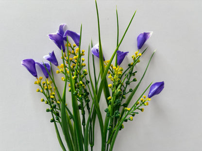 Full spread shot of an artificial purple small calla lily foliage bunch showing five individual wired stems fanning out with small purple calla lily flower heads, tall thin green grass stems and small bright yellow berry pick stems all arranged against a plain light grey background