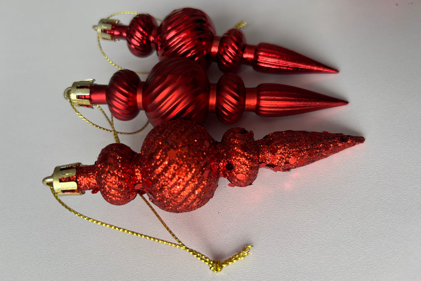 Three red Christmas spindle baubles stacked at an angle showing metallic and glittered finishes with ribbed spherical tops, beaded middles and elongated pointed drop bases with gold hanging hooks on a plain white background