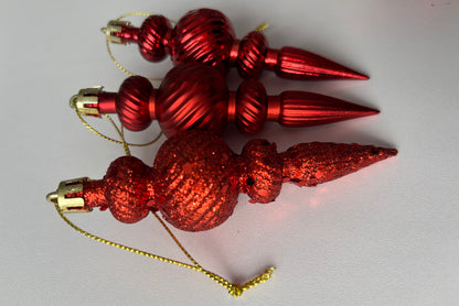 Three red Christmas spindle baubles stacked at an angle showing metallic and glittered finishes with ribbed spherical tops, beaded middles and elongated pointed drop bases with gold hanging hooks on a plain white background