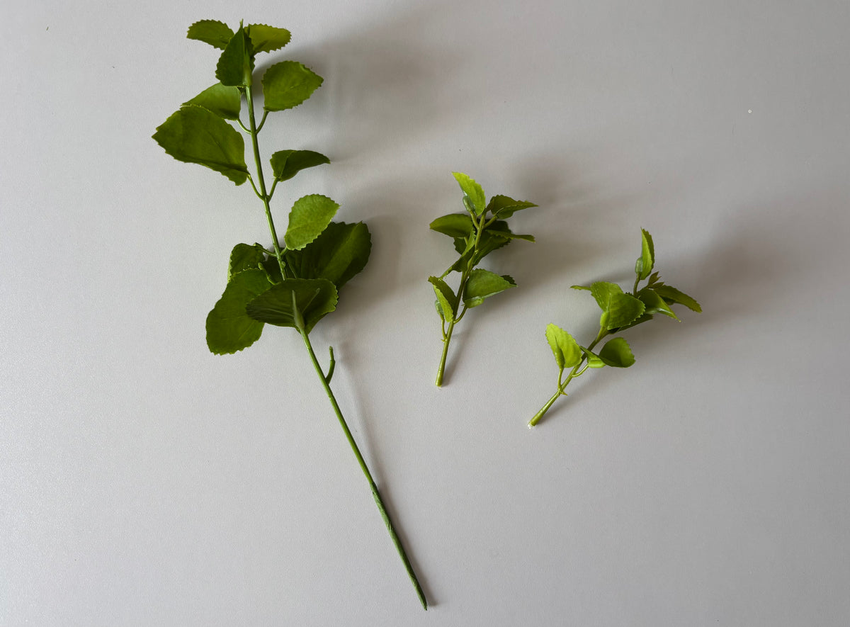 Flat lay shot of an artificial green patchouli leaf foliage spray showing the main spray stem laid diagonally on the left alongside two detached individual side branch stems laid separately to the right all displaying rounded serrated green fabric silk patchouli leaves against a plain light grey background showcasing the detachable stem feature