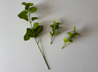 Flat lay shot of an artificial green patchouli leaf foliage spray showing the main spray stem laid diagonally on the left alongside two detached individual side branch stems laid separately to the right all displaying rounded serrated green fabric silk patchouli leaves against a plain light grey background showcasing the detachable stem feature