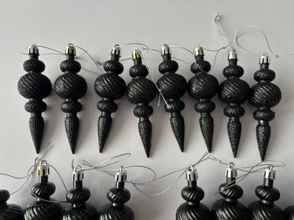 Flat lay of eight black glittered Christmas icicle finial baubles with ribbed spherical tops, beaded middles and elongated pointed drop bases with silver hanging strings arranged in two rows on a light grey background