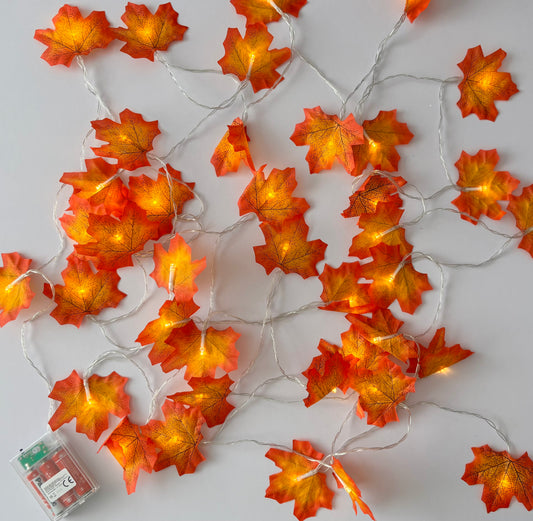 Overhead flat lay shot of an autumn maple leaf light up garland with 40 illuminated silk maple leaves in rich red and orange tones spread across a plain light grey background with battery box visible in the lower left corner