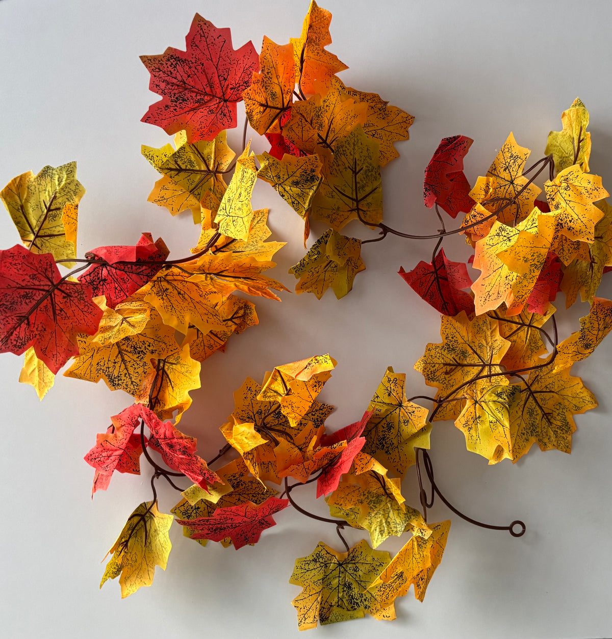 Overhead flat lay shot of an autumn maple leaf garland 180cm with multiple detachable double silk maple leaf sections in rich red, orange, yellow and green tones spread across a plain white background showing the full garland layout with brown plastic garland strand and two hanging hoops visible