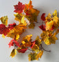Overhead flat lay shot of an autumn maple leaf garland 180cm with multiple detachable double silk maple leaf sections in rich red, orange, yellow and green tones spread across a plain white background showing the full garland layout with brown plastic garland strand and two hanging hoops visible