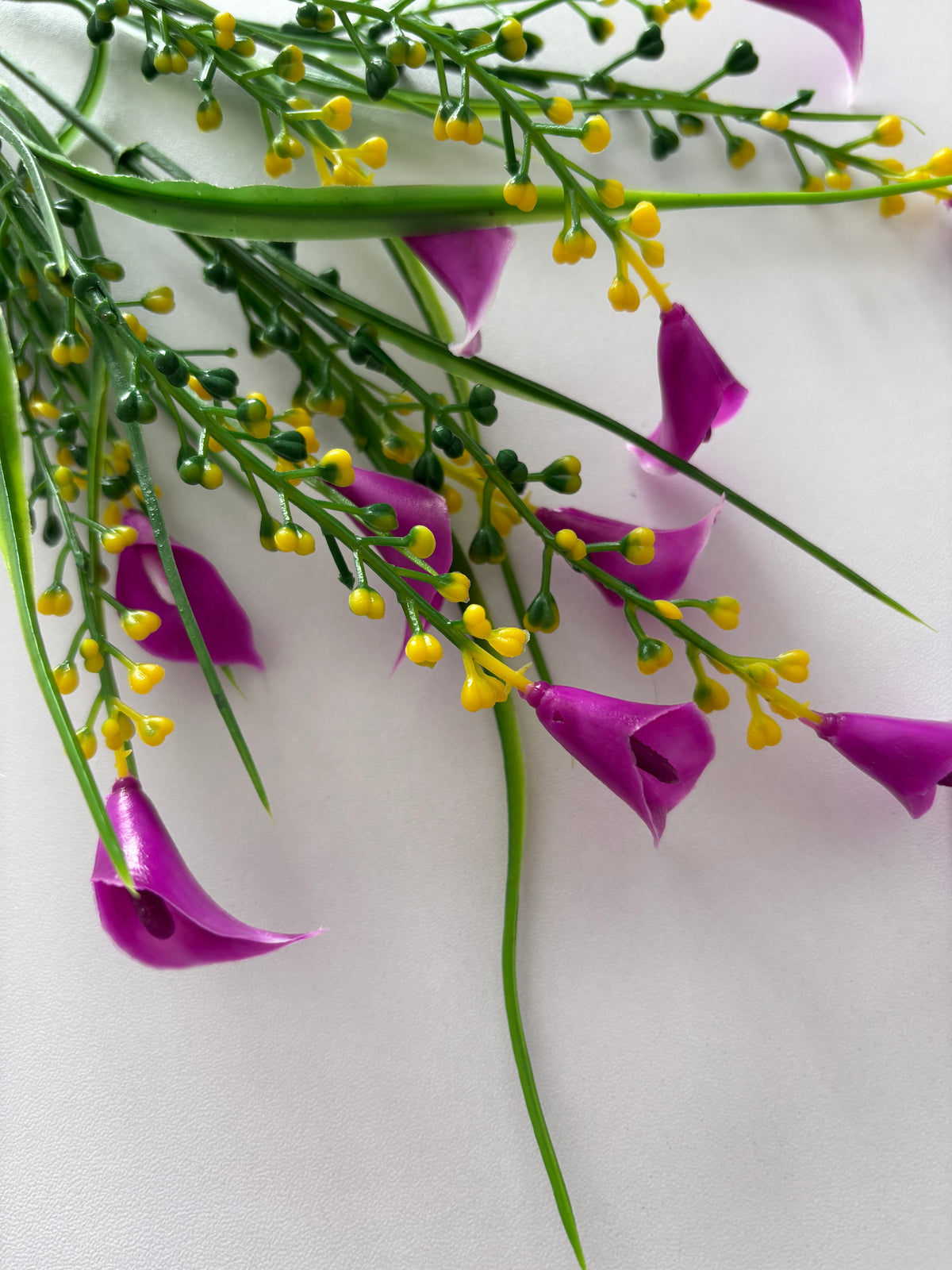 Extreme close up overhead macro shot of an artificial pink small calla lily foliage bunch showing multiple small bright magenta pink calla lily flower heads drooping naturally downward with beautiful white inner petal edges and dense clusters of bright yellow berry pick stems and thin green grass stems all visible against a plain white background