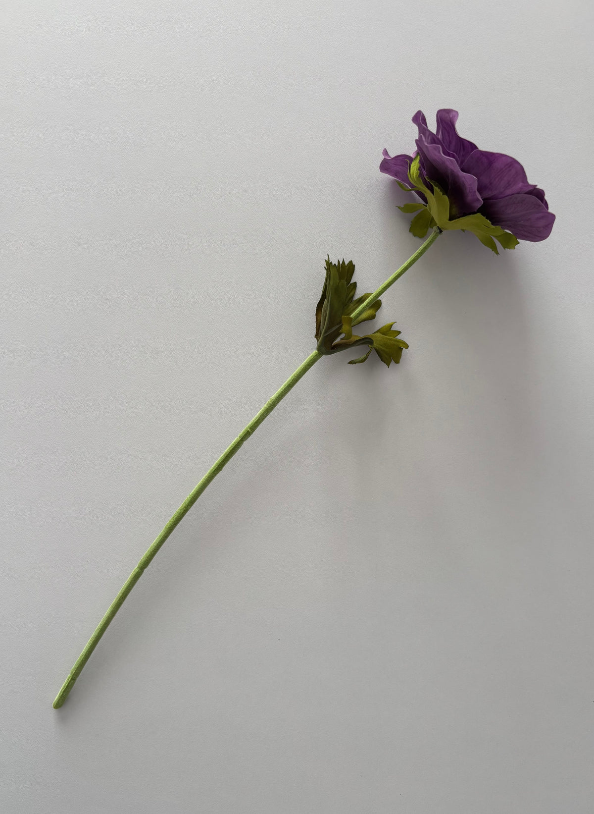 Full length flat lay shot of an artificial purple silk anemone single stem showing the complete 32cm stem with fabric wrapped wired stem, bright green detachable serrated foliage leaves and a closed purple anemone flower head at the top against a plain light grey background
