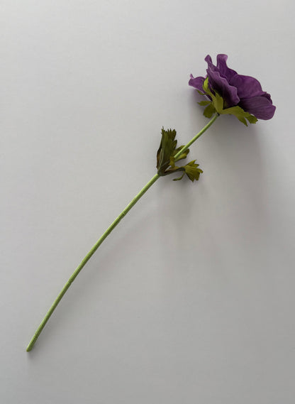 Full length flat lay shot of an artificial purple silk anemone single stem showing the complete 32cm stem with fabric wrapped wired stem, bright green detachable serrated foliage leaves and a closed purple anemone flower head at the top against a plain light grey background