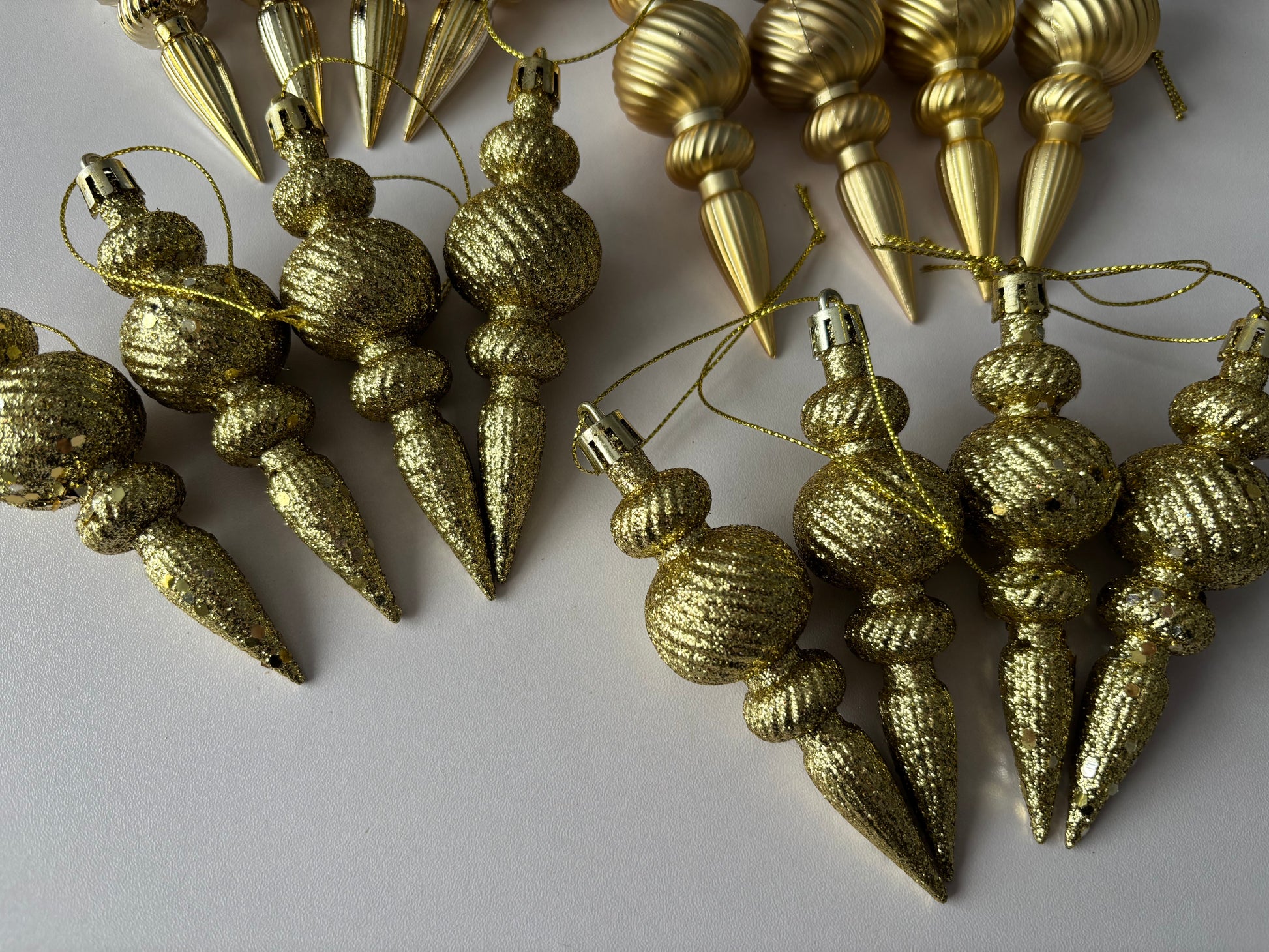 Close up of multiple gold Christmas spindle baubles showing glittered and metallic finishes with chunky glitter textured spherical tops, ribbed middles and elongated pointed drop bases with gold hanging strings on a plain white background