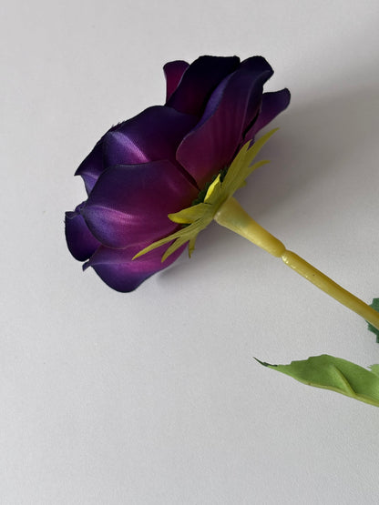Portrait shot of an artificial purple anemone single stem showing the closed partially open deep purple flower bud with dark purple petals fading to black at the edges bright yellow-green calyx and wired stem with dark green serrated foliage leaf on a plain light background