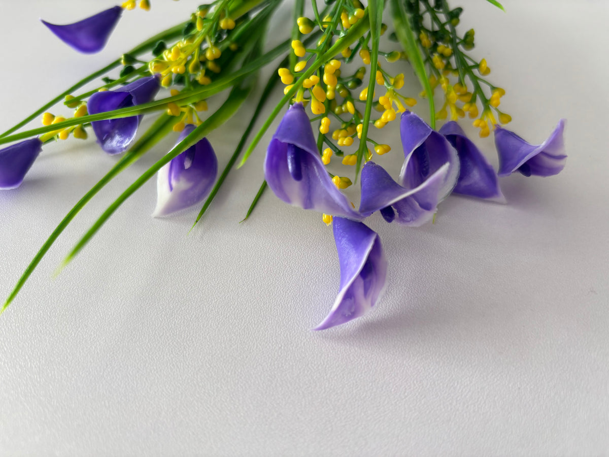 Extreme close up macro shot of an artificial purple small calla lily foliage bunch showing multiple small purple calla lily flower heads in the foreground with beautiful violet and white inner petal detail and soft petal texture with bright yellow berry pick clusters and green grass stems visible above against a plain white surface