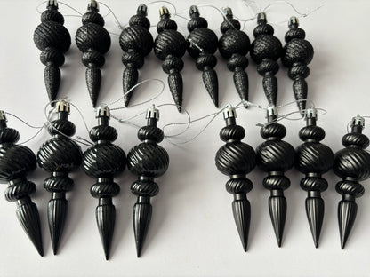 Flat lay of all 16 black Christmas icicle finial baubles in matte, metallic and glittered finishes arranged in two rows showing ribbed spherical tops, beaded middles and elongated pointed drop bases with silver hanging strings on a plain white background