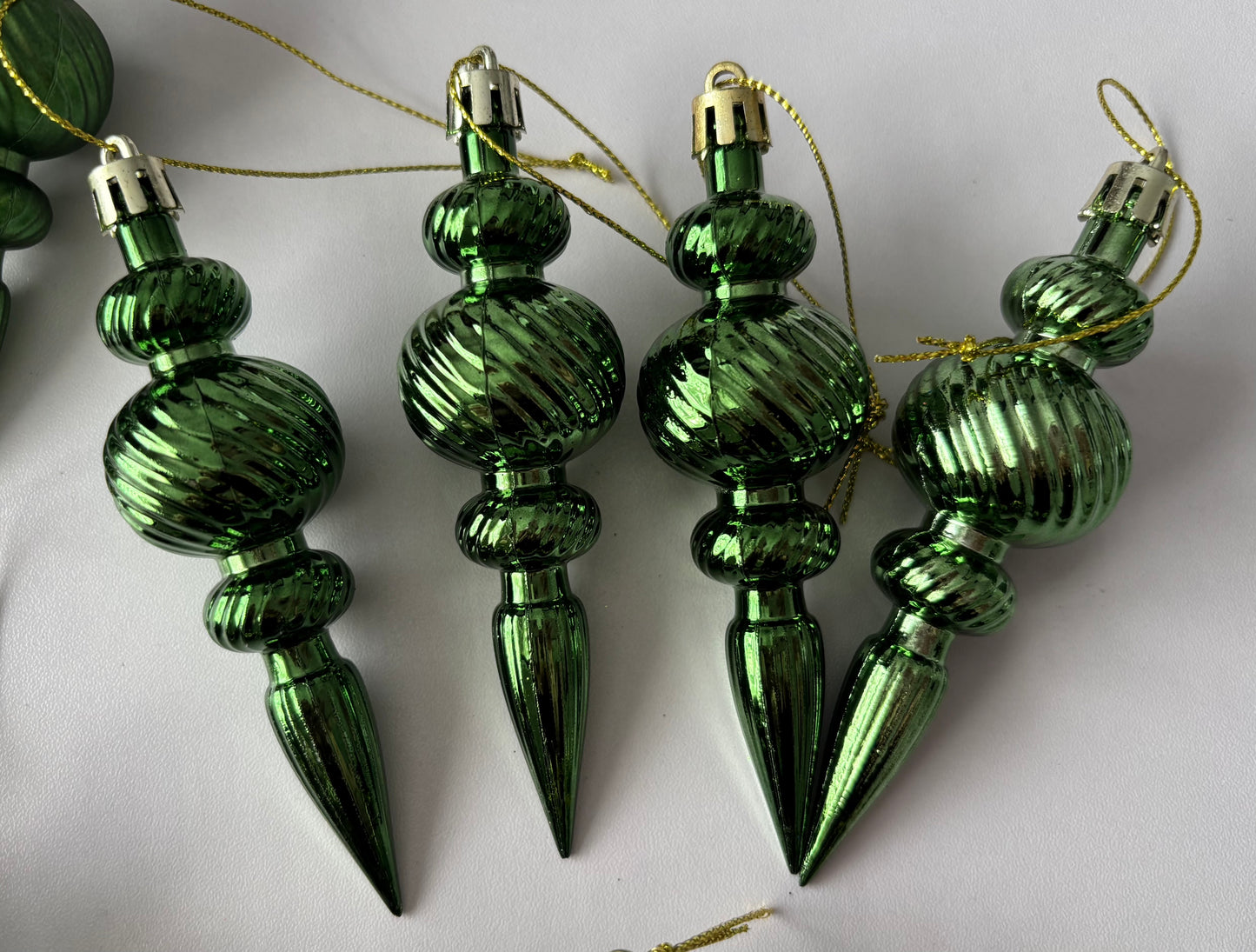 Close up of four green metallic Christmas spindle baubles with highly polished ribbed spherical tops, ribbed middles and elongated pointed drop bases with gold hanging hooks on a plain white background