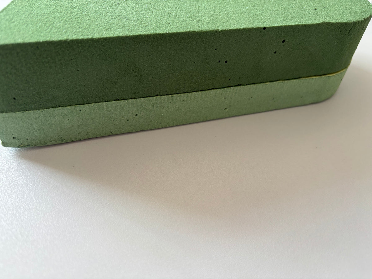 Low angle side profile shot of a green heart shaped oasis wet floral foam frame showing the solid firm base layer and two layer foam construction clearly with the lighter soft porous wet foam top layer and the darker solid green base visible against a plain white background