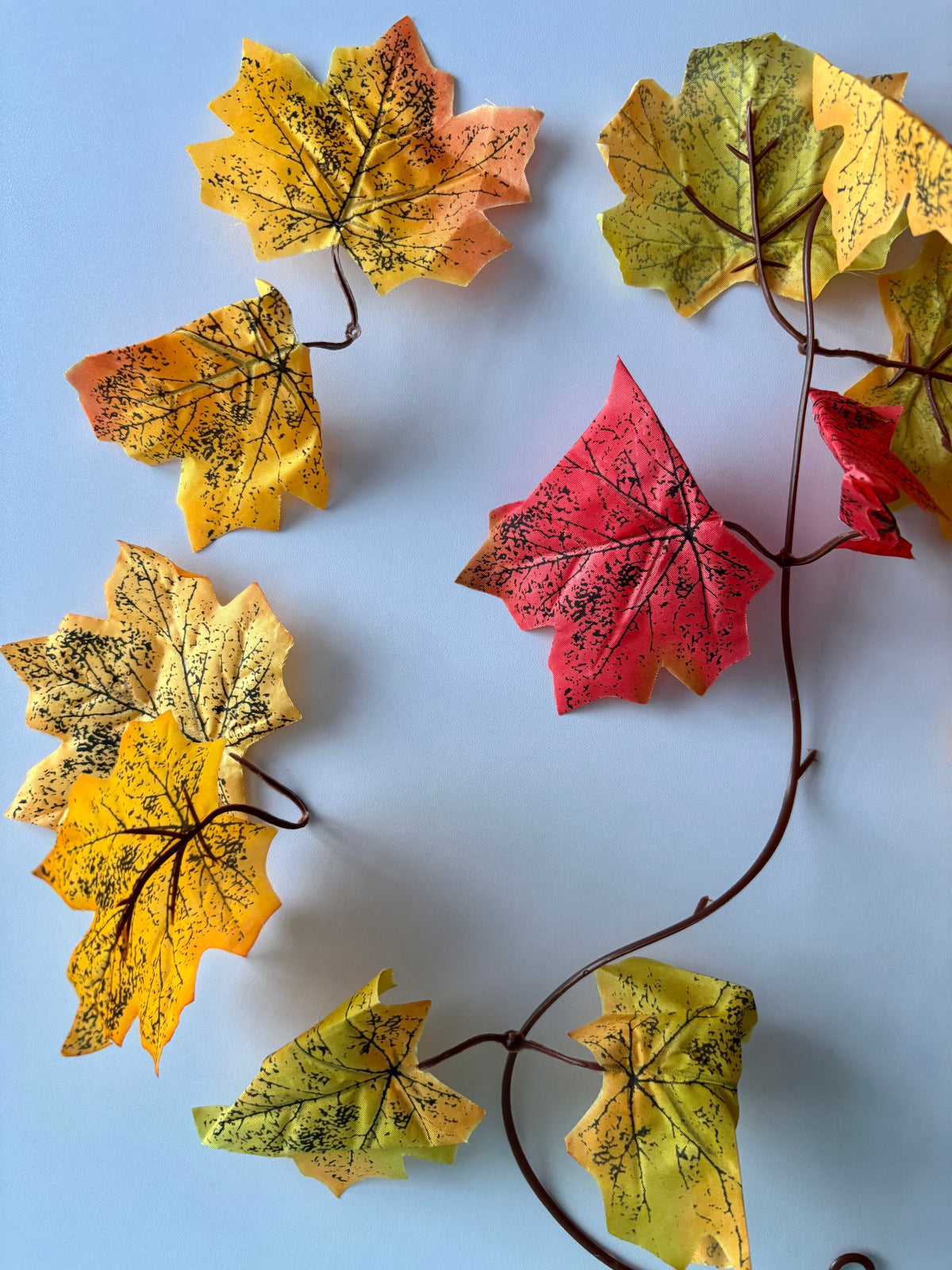 Flat lay shot of an autumn maple leaf garland 180cm showing individual detachable double silk maple leaf sections spread out along the brown plastic garland strand in yellow, orange, red and green tones with detailed black leaf vein markings against a plain light blue background