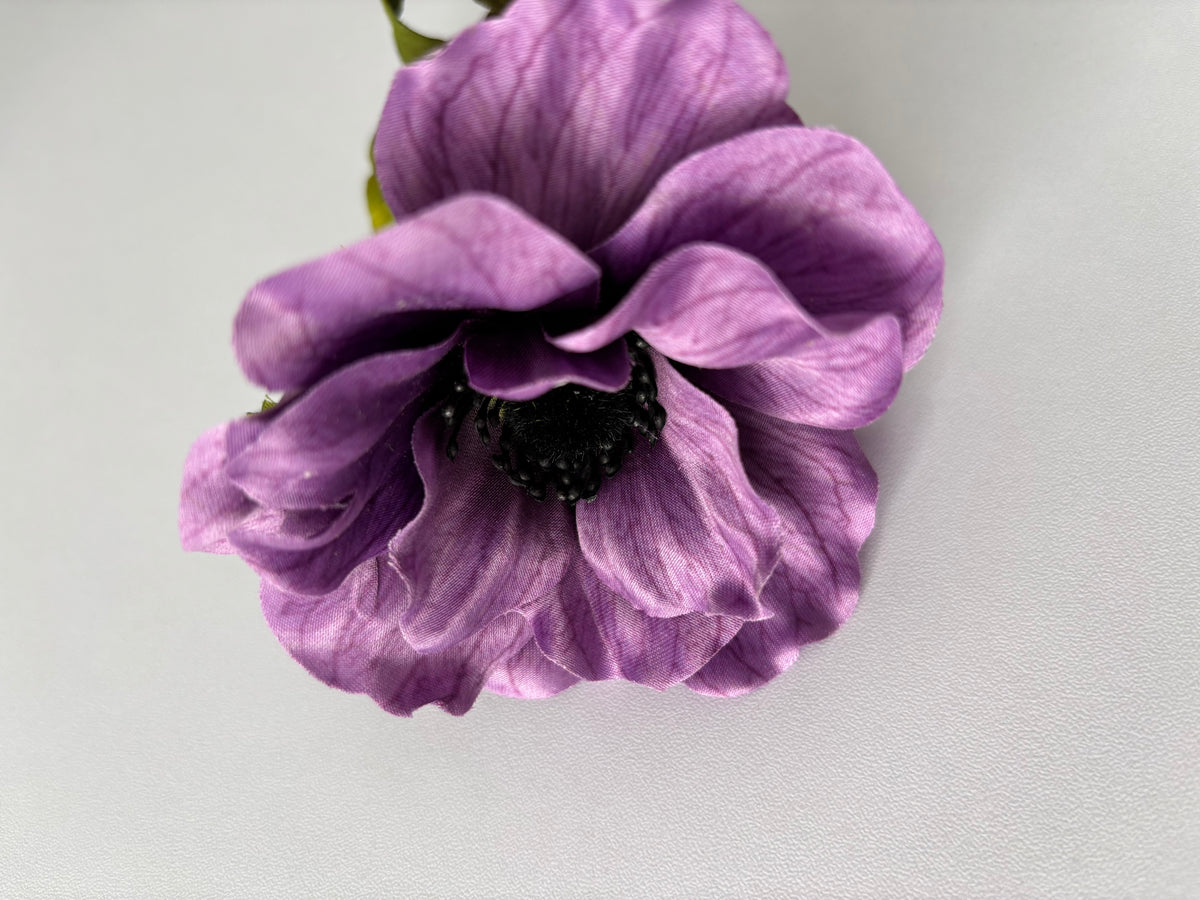 Slightly angled overhead shot of a large artificial purple silk anemone flower head showing beautifully layered open ruffled petals with fine vein detail throughout and a dramatic raised dark black seed centre with tiny black stamens against a plain light background with green foliage just visible behind