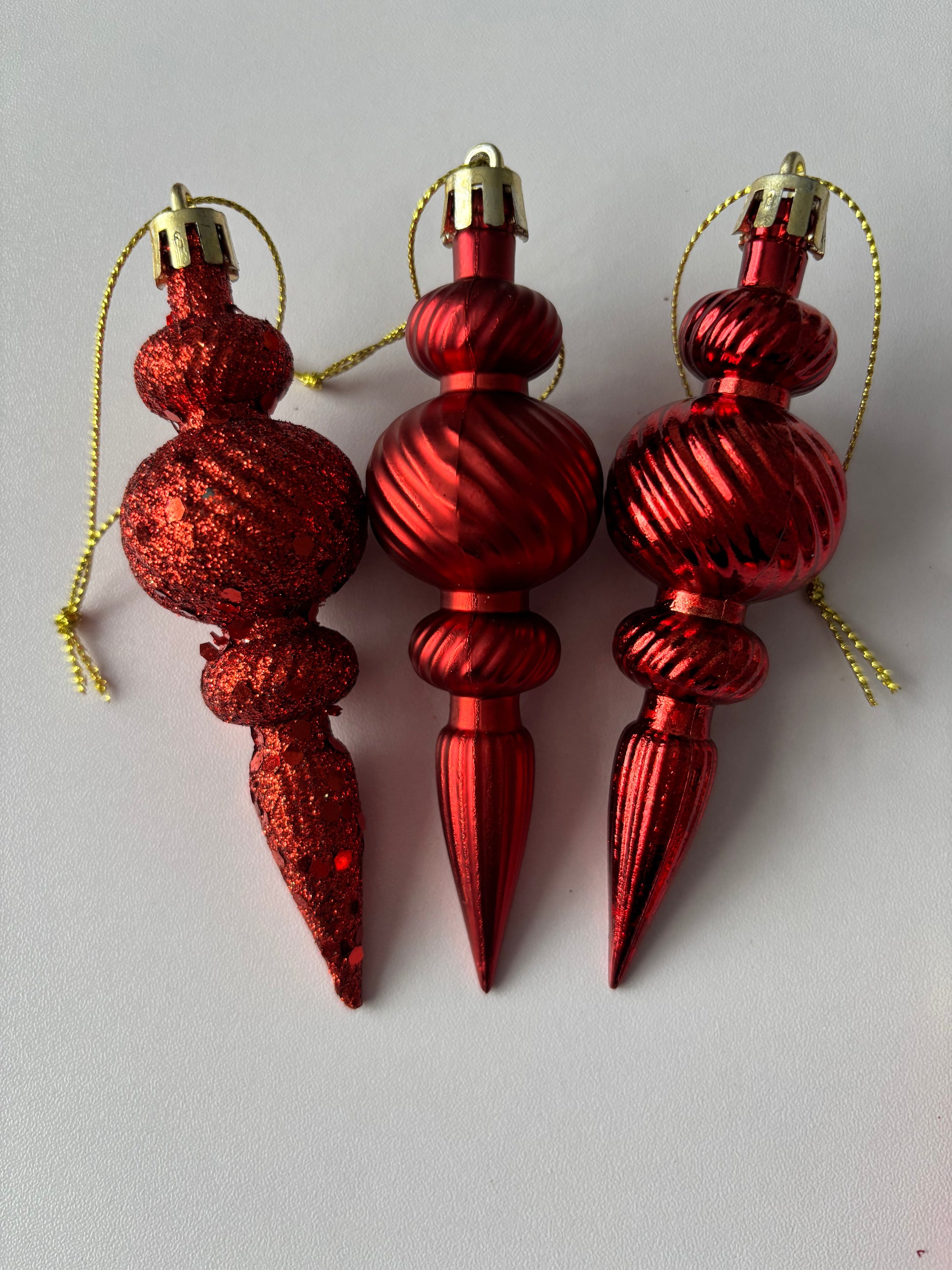 Three red Christmas spindle baubles side by side showing glittered, metallic and matte finishes with ribbed spherical tops, beaded middles and elongated pointed drop bases with gold hanging hooks on a plain white background