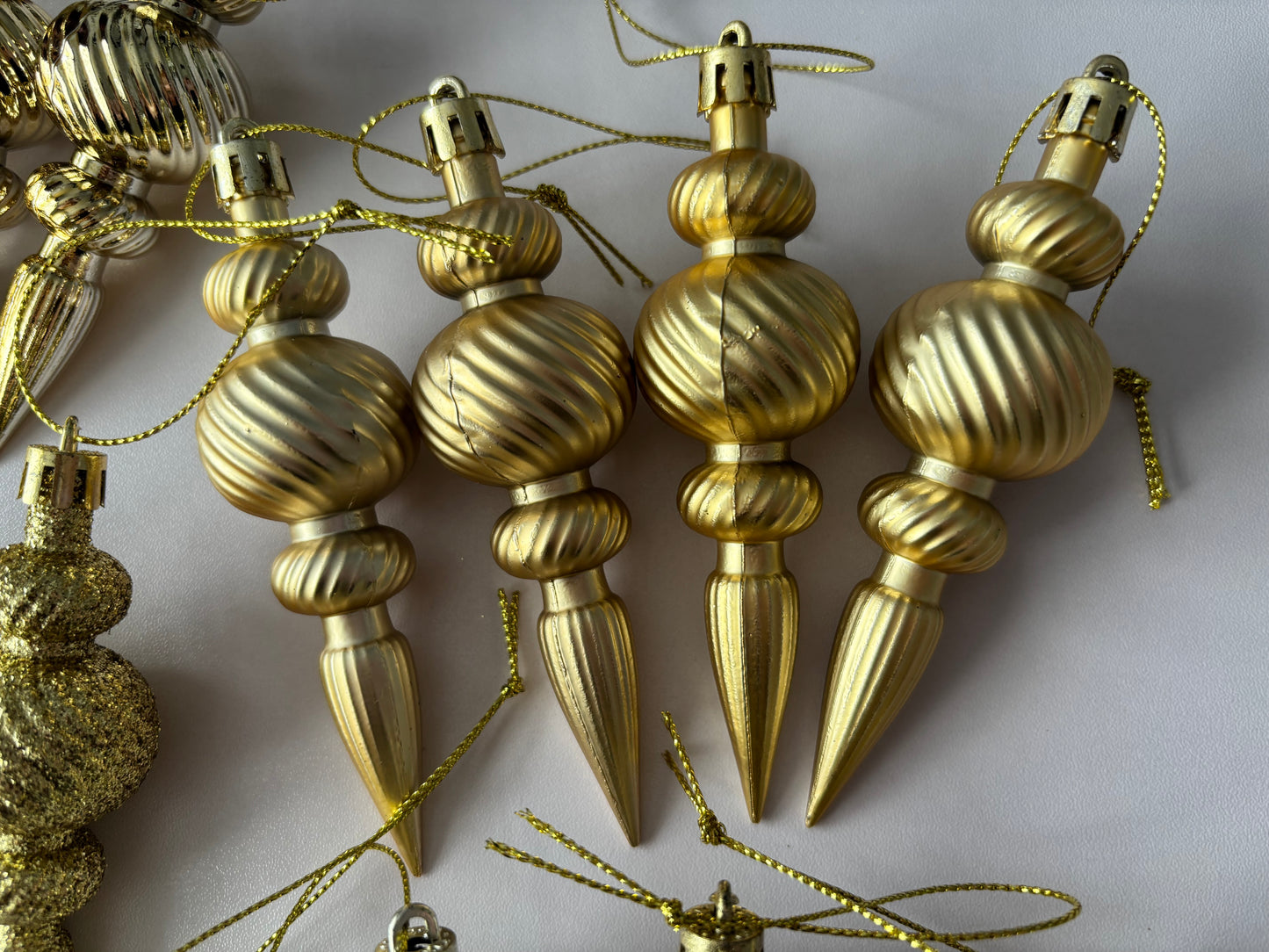 Close up of four gold metallic Christmas spindle baubles with highly polished ribbed spherical tops, twisted ribbed middles and elongated pointed drop bases with gold hanging hooks on a plain light grey background
