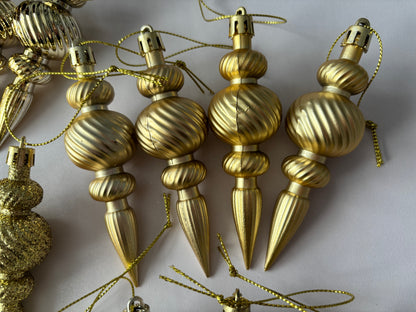 Close up of four gold metallic Christmas spindle baubles with highly polished ribbed spherical tops, twisted ribbed middles and elongated pointed drop bases with gold hanging hooks on a plain light grey background