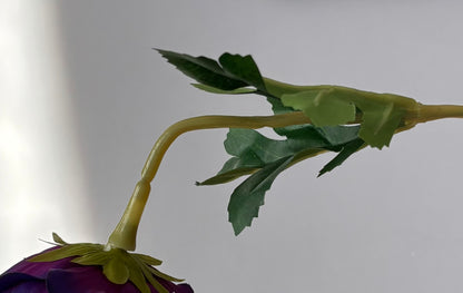 Extreme close up shot of the yellow-green wired stem and dark green serrated foliage leaves of an artificial purple anemone single stem with the deep purple flower head partially visible at the bottom on a plain light grey background