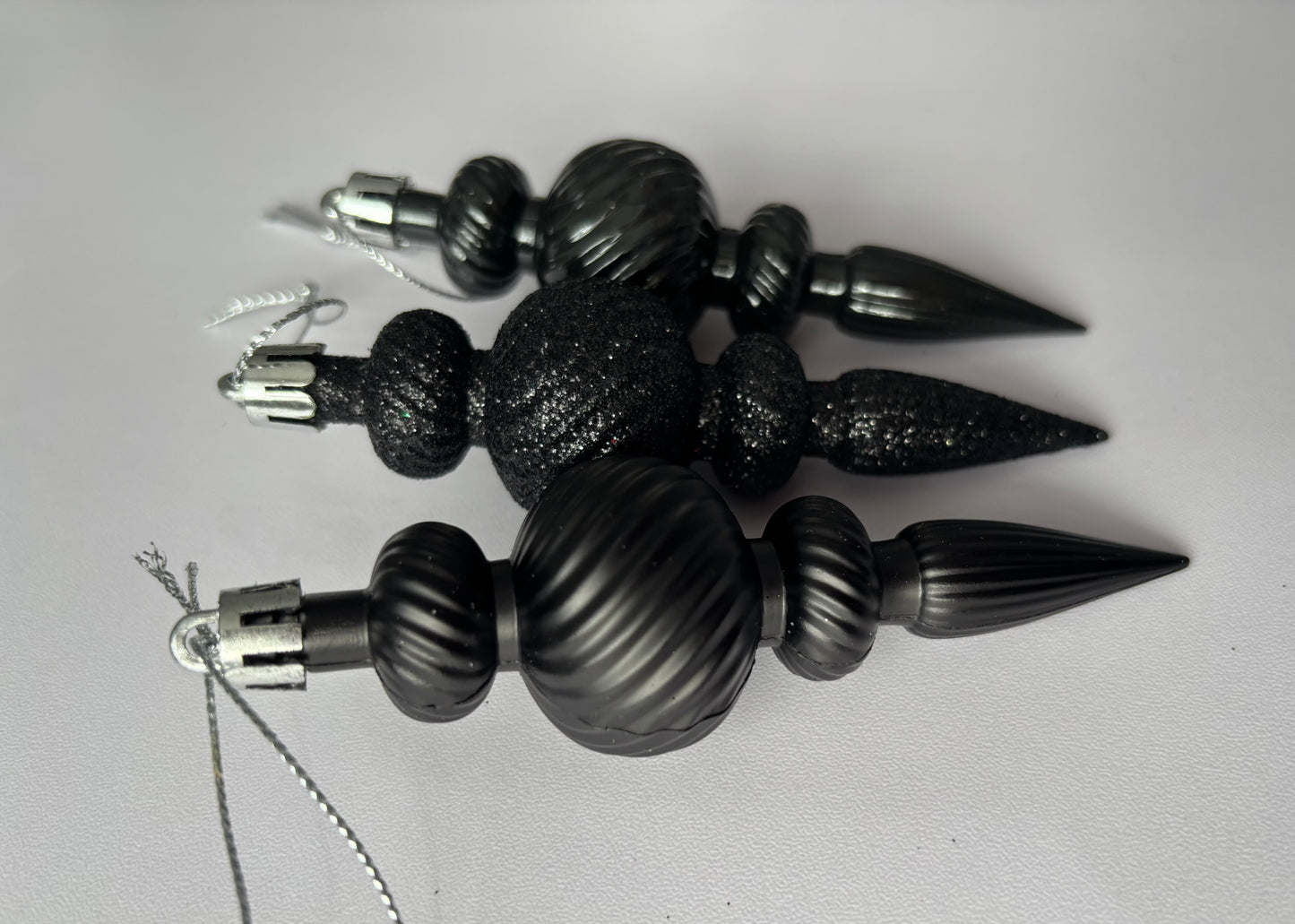 Close up of three black Christmas icicle finial baubles showing matte, glittered and metallic finishes with ribbed spherical tops, twisted ribbed middles and pointed spike bases with silver hanging hooks on a light grey background