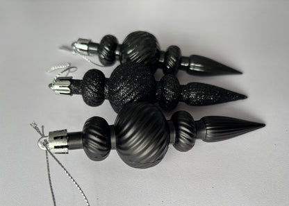 Close up of three black Christmas icicle finial baubles showing matte, glittered and metallic finishes with ribbed spherical tops, twisted ribbed middles and pointed spike bases with silver hanging hooks on a light grey background