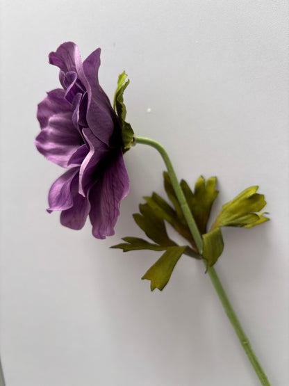 Side profile shot of an artificial purple silk anemone single stem showing the open flower head with layered ruffled purple petals and dark black centre viewed from the side with two large bright green deeply serrated detachable foliage leaves spread wide and the fabric wrapped green stem visible below against a plain light background