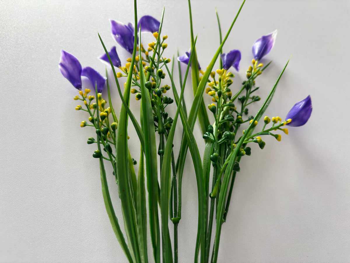 Diagonal flat lay shot of an artificial purple small calla lily foliage bunch laid across a plain light background showing the full stem length with small purple calla lily flower heads drooping naturally at the tips, clusters of bright yellow berry picks and tall thin green grass stems all fanning out from the single bottom stem
