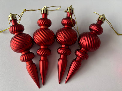 Close up of four red metallic Christmas spindle baubles with highly polished ribbed spherical tops, twisted ribbed middles and elongated pointed drop bases with gold hanging hooks arranged side by side on a plain white background