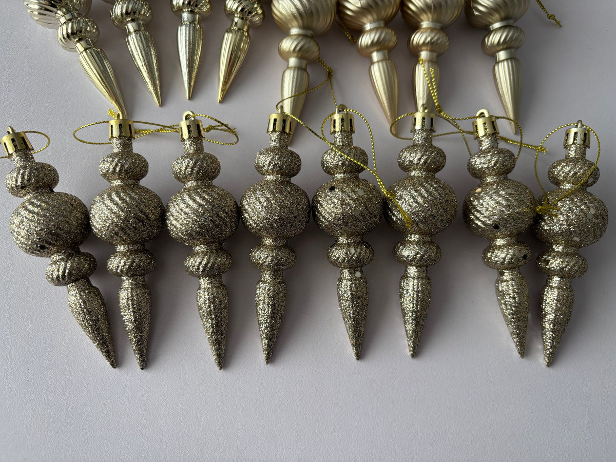 Close up overhead flat lay of eight champagne gold glittered finish Christmas spindle baubles with chunky glitter textured ribbed spherical tops, twisted middles and elongated pointed drop bases with gold hanging strings arranged on a plain light grey background