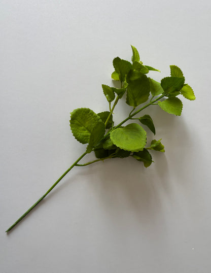 Diagonal flat lay shot of an artificial green patchouli leaf foliage spray showing the full stem length with a main wired plastic stem, two branch stems and multiple variety-sized rounded serrated green fabric silk leaves arranged naturally against a plain light grey background