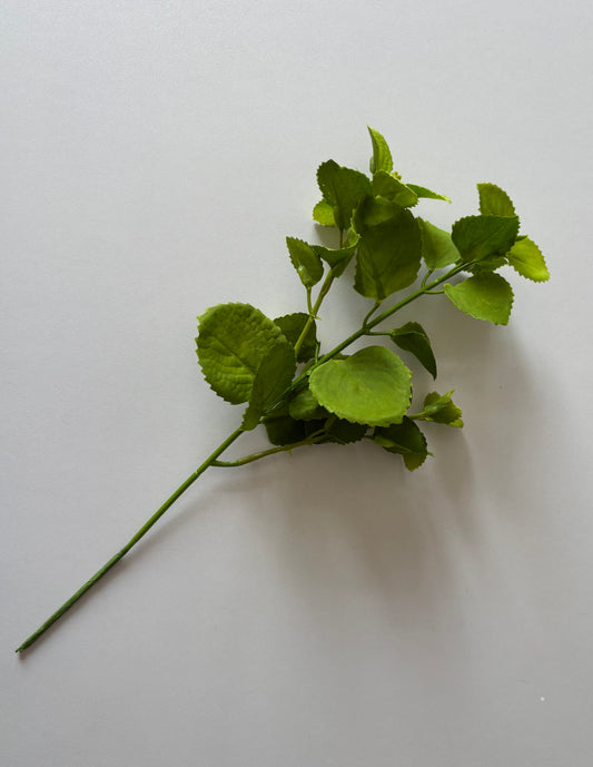Diagonal flat lay shot of an artificial green patchouli leaf foliage spray showing the full stem length with a main wired plastic stem, two branch stems and multiple variety-sized rounded serrated green fabric silk leaves arranged naturally against a plain light grey background