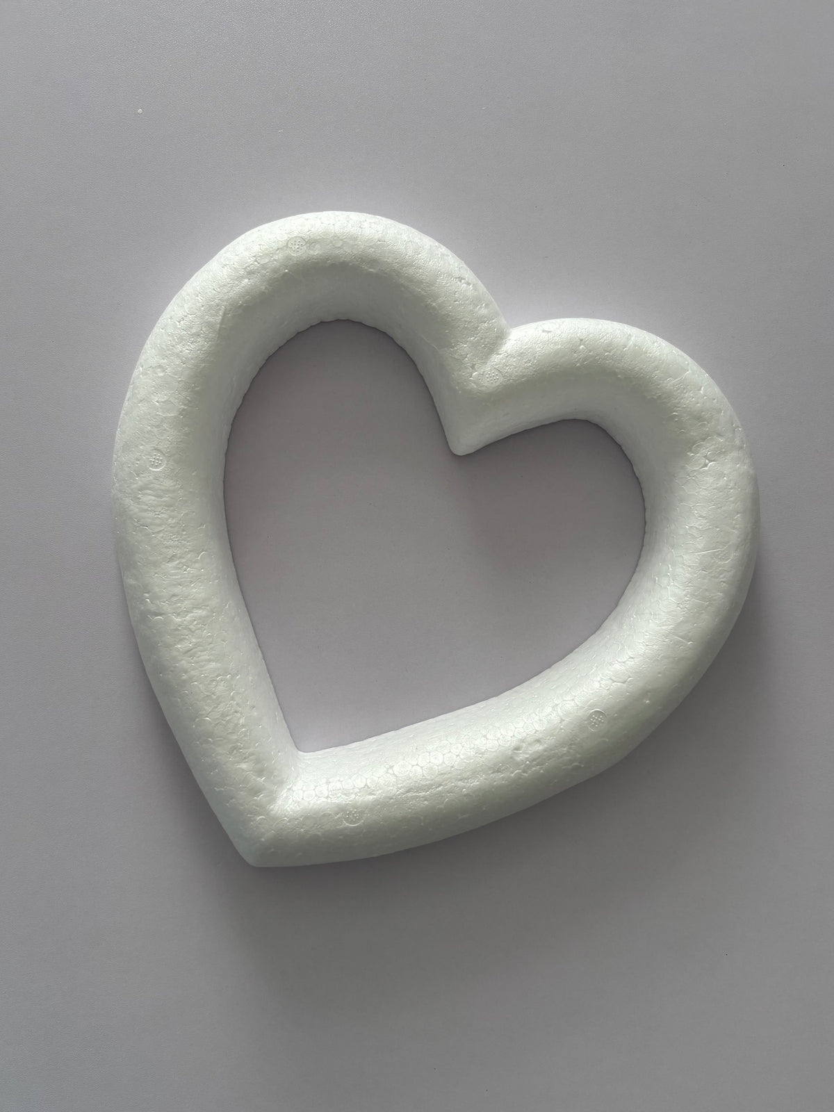Overhead flat lay shot of a white polystyrene heart wreath frame 28cm showing the full hollow heart ring shape with thick textured polystyrene border against a plain neutral grey background