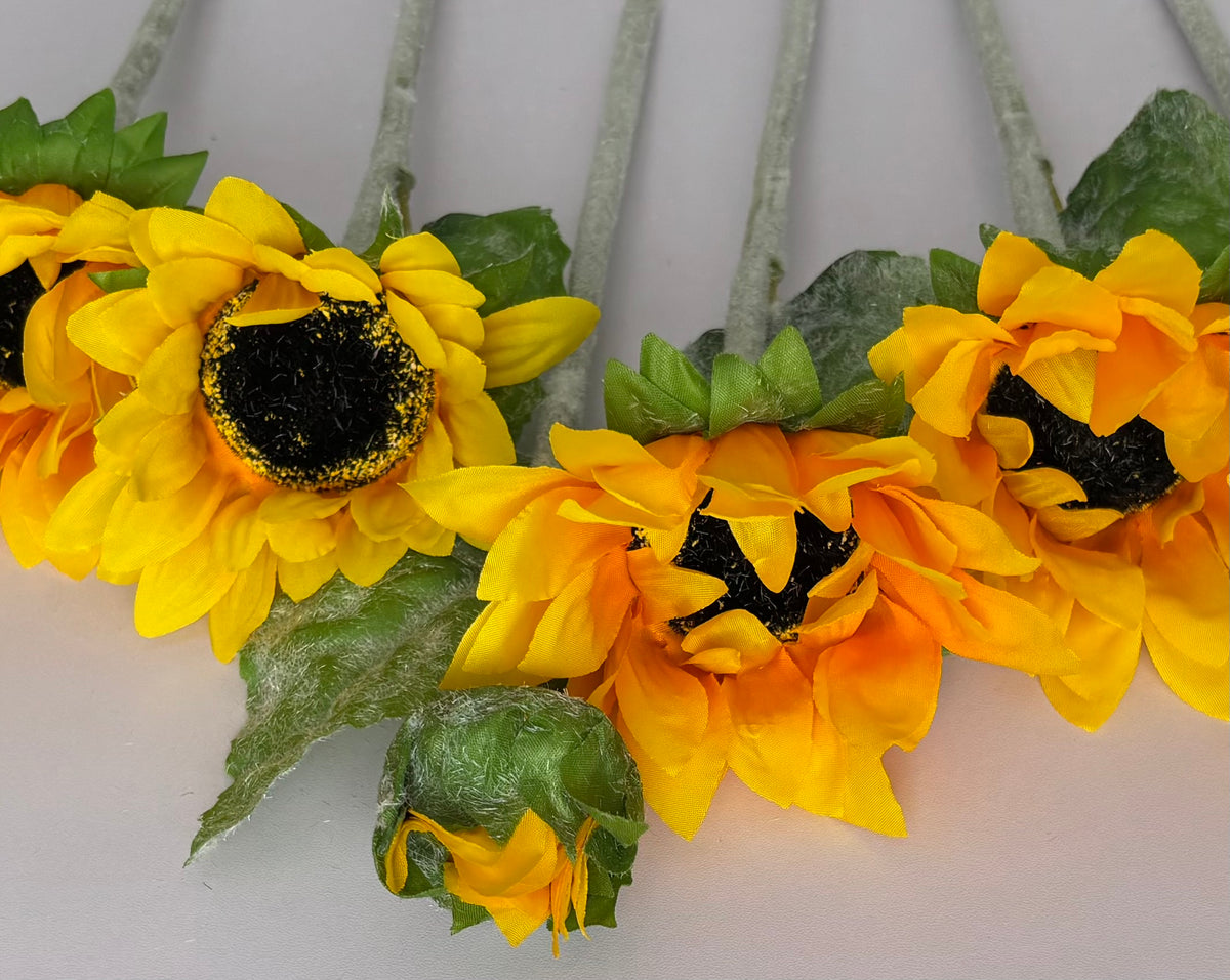 Close up shot of three artificial sunflower heads laid horizontally showing the mixed size golden yellow and orange petals with dark black textured centres one glittery centre and lush dark green foliage with glittery silver fuzzy stems on a plain light grey background