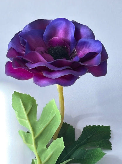 Portrait shot of an artificial purple anemone single stem stood upright showing the full stem with stunning layered deep purple and magenta flower head dark green serrated foliage leaves and yellow-green wired stem on a plain light grey background