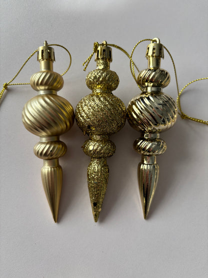 Close up of three gold Christmas spindle baubles side by side showing metallic, glittered and matte finishes with ribbed spherical tops, beaded middles and elongated pointed drop bases with gold hanging strings on a plain light grey background